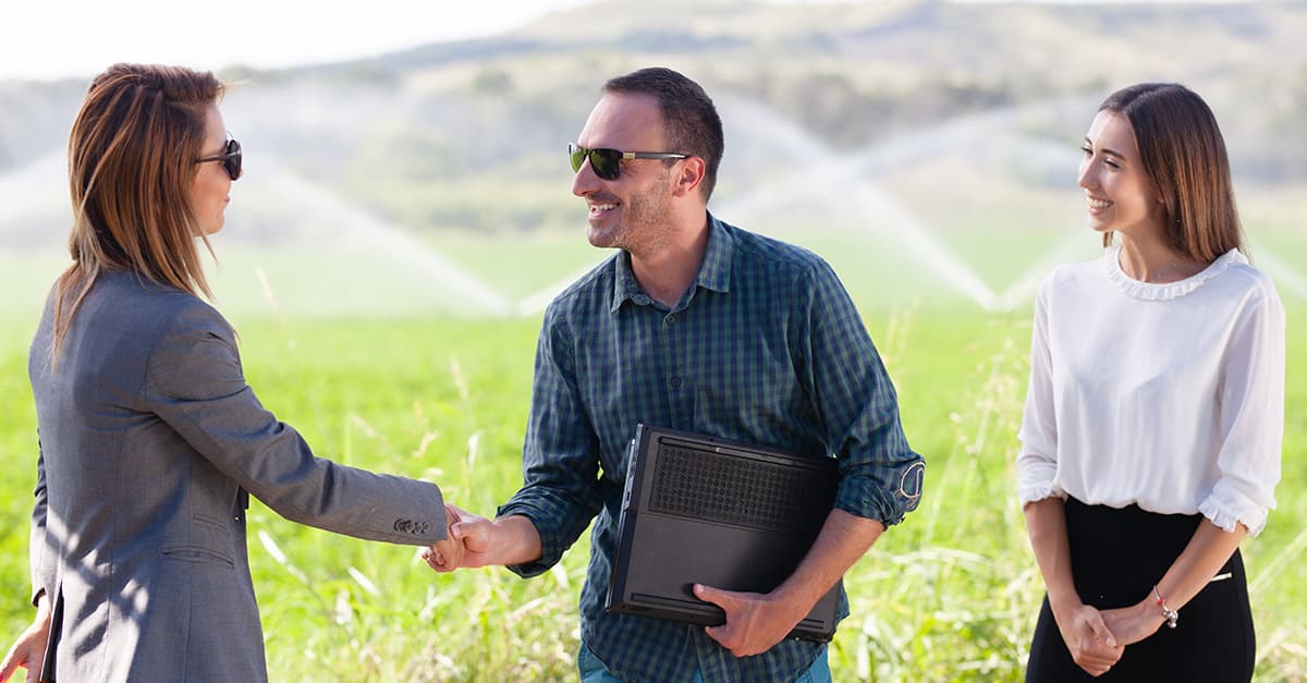 Man shaking woman's hand over business deal with another businesswoman watching, in a field with sprinklers running.