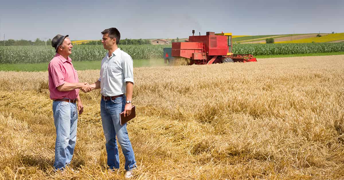 Two men shaking hands in a field while a combine harvests wheat.