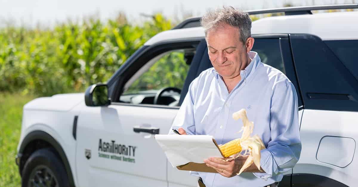 Horacio Buscaglia wearing a light blue button-up shirt stands in front of a white AgriThority® vehicle in a cornfield. He is holding a clipboard and an ear of corn while writing notes, with a focused expression on his face. This image supports the article on international market expansion.