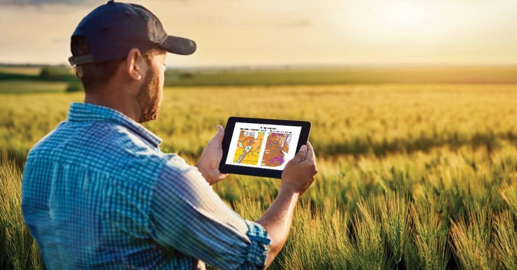 A farmer in a plaid shirt and cap stands in a wheat field at sunset, holding a tablet displaying two colorful maps. The maps show agricultural data, such as yield or soil analysis, highlighting precision farming techniques. The golden field stretches into the distance under a soft, glowing sky.