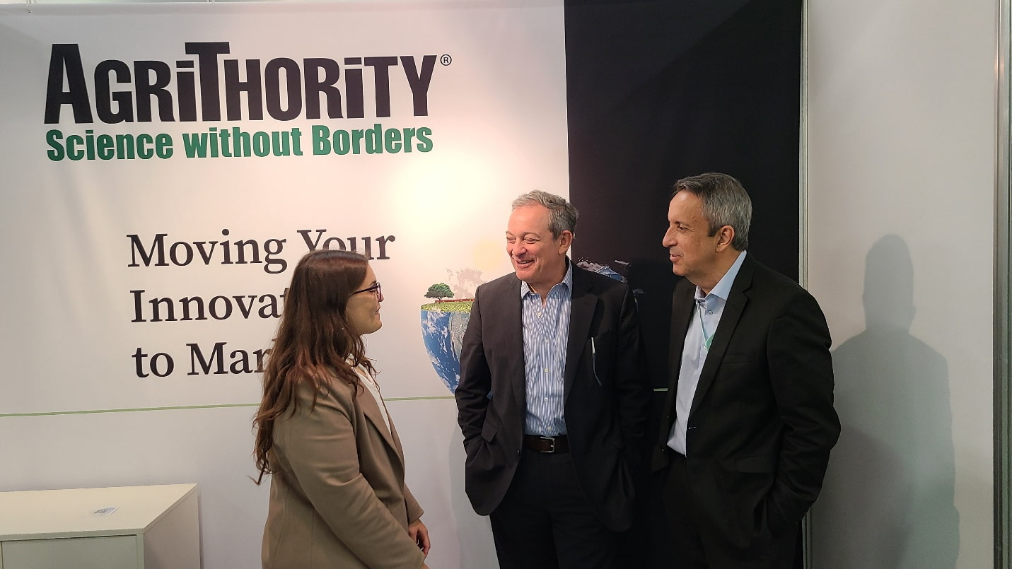 Three AgriThority® team members in business attire discussing at the AgriThority® booth, featuring the slogan "Science without Borders" and "Moving Your Innovation to Market," with a backdrop of a global map and greenery, ideal for agriculture and innovation insights.