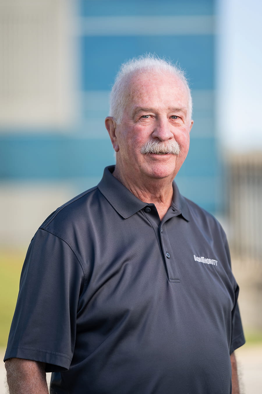 Headshot of Jerry Duff, Founder & President of AgriThority® with white hair and a mustache, wearing a dark gray polo shirt with the ‘AgriThority®’ logo, standing outdoors in front of a modern building with blue and white panels.