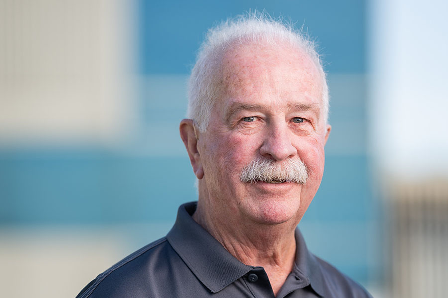Headshot of Jerry Duff, Founder & President of AgriThority® with white hair and a mustache, wearing a dark gray polo shirt with the ‘AgriThority®’ logo, standing outdoors in front of a modern building with blue and white panels.